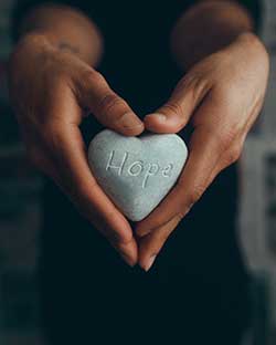 Two hands holding a heart-shaped rock with the word Hope engraved in it.